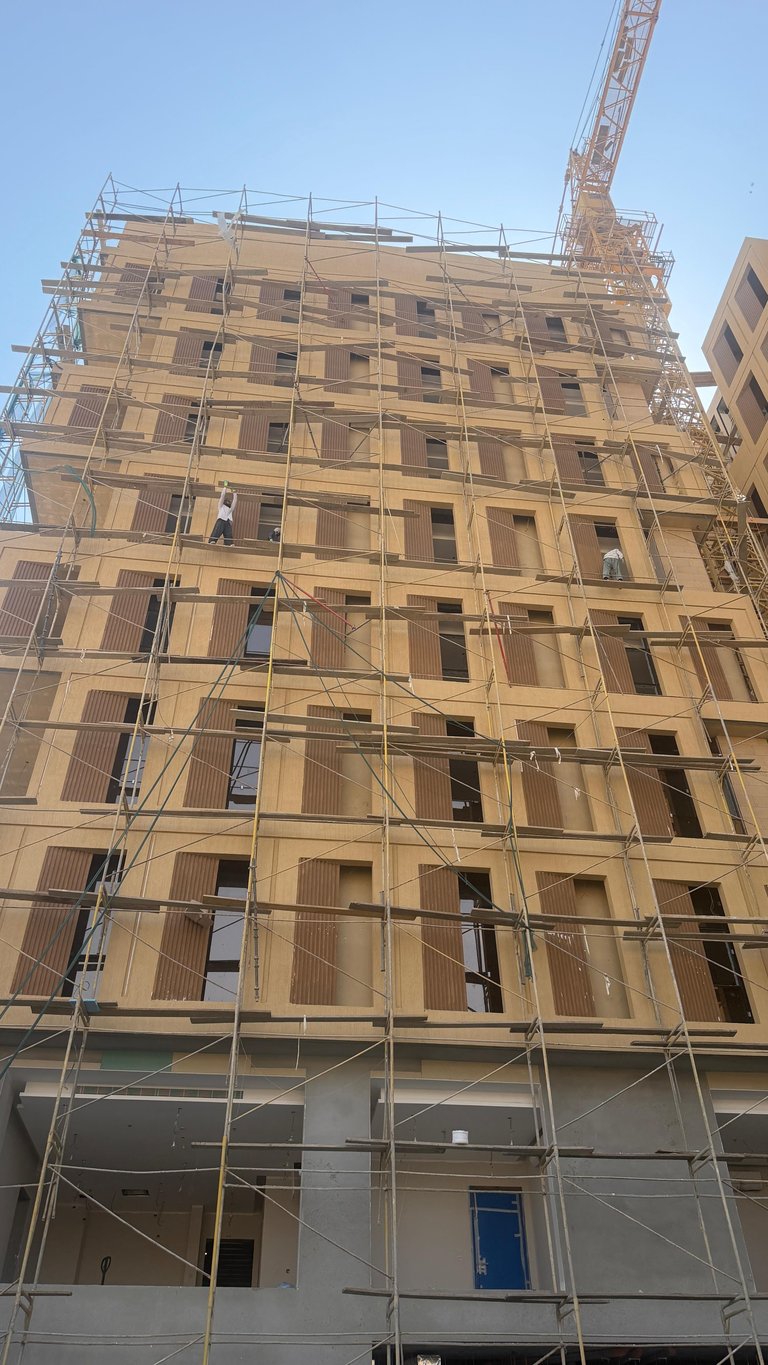 Multi-story residential building under construction with metal scaffolding, partially completed facade, and construction crane visible against clear blue sky